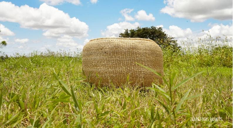 Puede incluir: Una cesta grande, redonda y tejida de forma natural se encuentra en un campo de hierba verde. La cesta est&aacute; hecha de materiales naturales y tiene un color marr&oacute;n claro. El cielo es azul con nubes blancas en el fondo.