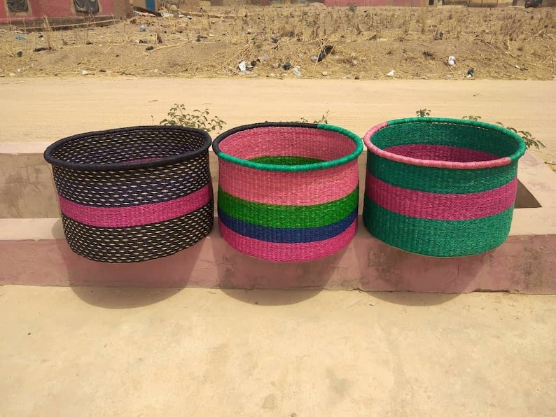 May include: Three woven baskets in a row. The baskets are cylindrical with colorful horizontal stripes. The first basket is black with a pink stripe. The second basket is pink, green, and blue. The third basket is green and pink.