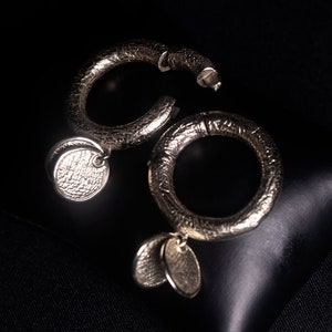 Puede incluir: Un par de pendientes de aro de plata con superficies texturizadas y colgantes con forma de moneda.