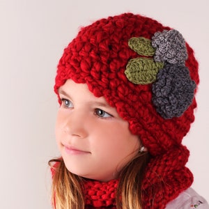 May include: A young girl wearing a red knitted hat with green and grey flower embellishments and a matching red knitted scarf.