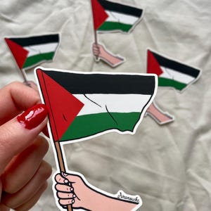 May include: A sticker of a hand holding a Palestinian flag. The flag is red, white, and green with a black triangle in the upper hoist corner. The sticker is signed "Siramouche".
