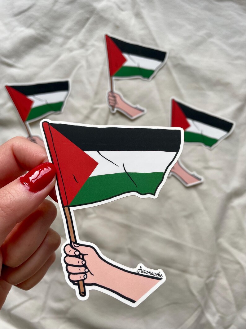 May include: A sticker of a hand holding a Palestinian flag. The flag is red, white, and green with a black triangle in the upper hoist corner. The sticker is signed "Siramouche".