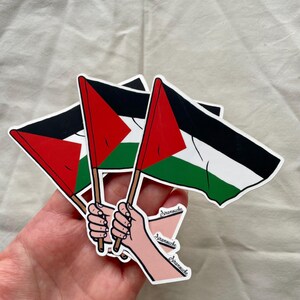 May include: Three Palestinian flags are held up by three hands. The flags are black, white, red, and green. The hands are drawn in a cartoon style and have the word "Dramouche" written on them.