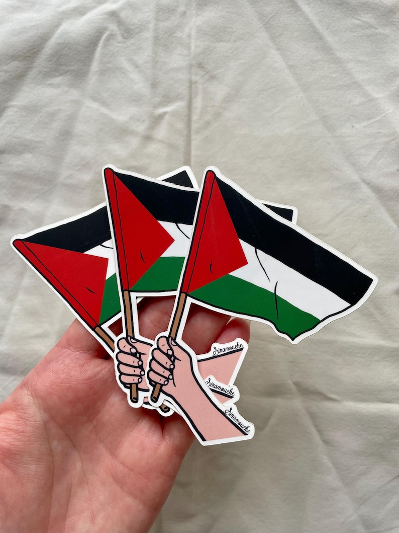 May include: Three Palestinian flags are held up by three hands. The flags are black, white, red, and green. The hands are drawn in a cartoon style and have the word "Dramouche" written on them.