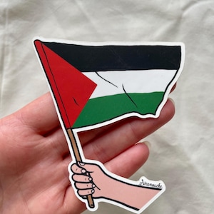 May include: A sticker of a hand holding a Palestinian flag. The flag is black, white, red, and green. The sticker is signed "Siramouche".