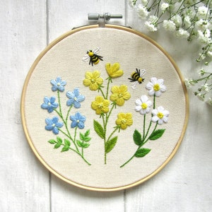 May include: Embroidered floral hoop art featuring blue, yellow, and white flowers with green leaves. Two bees are embroidered above the flowers. The embroidery is set in a wooden hoop, perfect for wall decor.