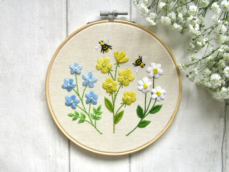 May include: Embroidered floral hoop art featuring blue, yellow, and white flowers with green leaves. Two bees are embroidered above the flowers. The embroidery is set in a wooden hoop, perfect for wall decor.