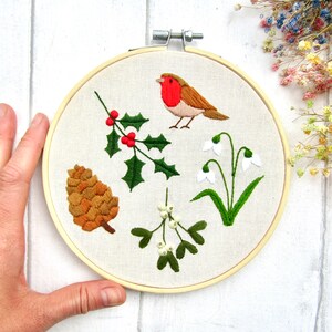 May include: Embroidered hoop art featuring a robin, holly with red berries, a pinecone, mistletoe, and snowdrops. The design is stitched on a cream-colored fabric, set in a wooden hoop. A charming, nature-themed piece.