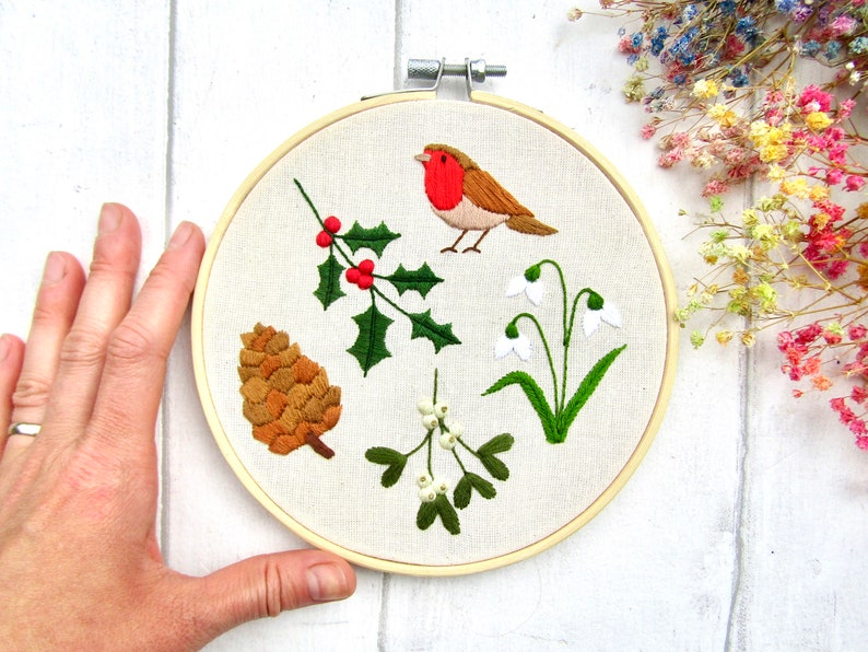 May include: Embroidered hoop art featuring a robin, holly with red berries, a pinecone, mistletoe, and snowdrops. The design is stitched on a cream-colored fabric, set in a wooden hoop. A charming, nature-themed piece.