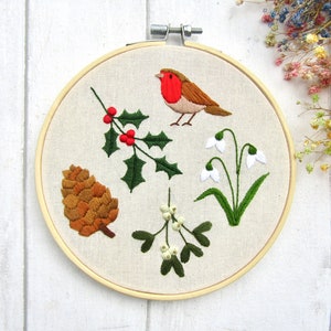 May include: Embroidered hoop art featuring a robin, holly, snowdrops, a pinecone, and mistletoe. The bird has a red breast and brown feathers. The holly has green leaves and red berries. The snowdrops have white petals and green stems.