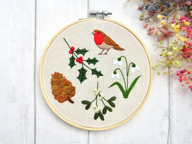 May include: Embroidered hoop art featuring a robin, holly, snowdrops, a pinecone, and mistletoe. The bird has a red breast and brown feathers. The holly has green leaves and red berries. The snowdrops have white petals and green stems.