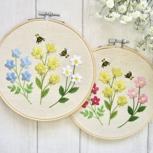 May include: Two embroidered floral hoops. One features blue, yellow, and white flowers with bees. The other has yellow, pink, and red flowers with bees. Both are set in wooden hoops with a cream-colored fabric background.
