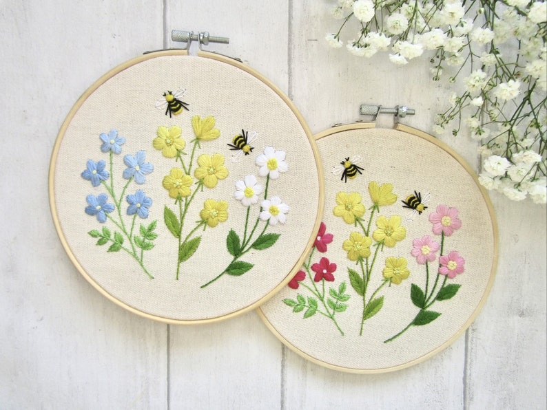 May include: Two embroidered floral hoops. One features blue, yellow, and white flowers with bees. The other has yellow, pink, and red flowers with bees. Both are set in wooden hoops with a cream-colored fabric background.