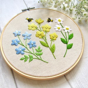 May include: Embroidered floral hoop art featuring blue, yellow, and white flowers with green leaves and two embroidered bees. The flowers are arranged on a beige fabric stretched within a wooden hoop. A charming, handmade piece.