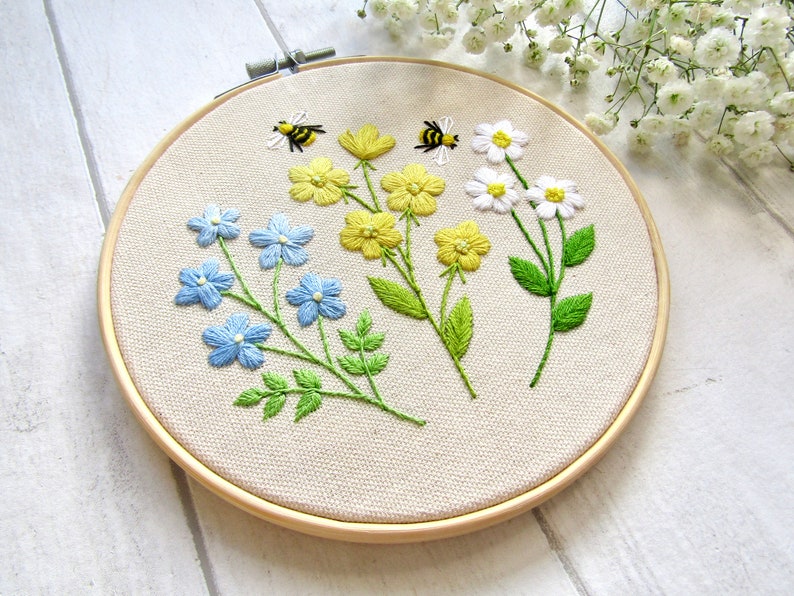 May include: Embroidered floral hoop art featuring blue, yellow, and white flowers with green leaves and two embroidered bees. The flowers are arranged on a beige fabric stretched within a wooden hoop. A charming, handmade piece.