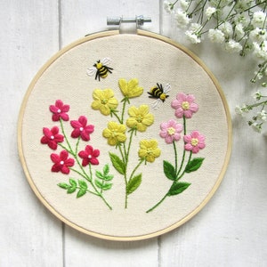 May include: Embroidered floral hoop art featuring three types of flowers in shades of pink and yellow, with green leaves and two embroidered bees. The artwork is set against a cream-colored fabric, mounted in a wooden hoop.
