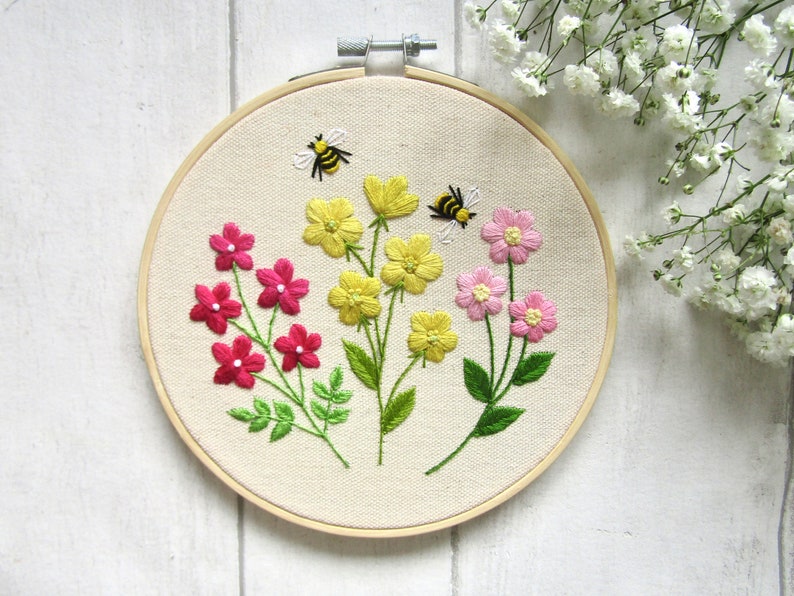 May include: Embroidered floral hoop art featuring three types of flowers in shades of pink and yellow, with green leaves and two embroidered bees. The artwork is set against a cream-colored fabric, mounted in a wooden hoop.