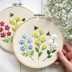 May include: Two round embroidery hoops with floral designs. One hoop features red flowers, the other blue and yellow flowers. Both have embroidered bees. The hoops are set against a white background with white flowers.