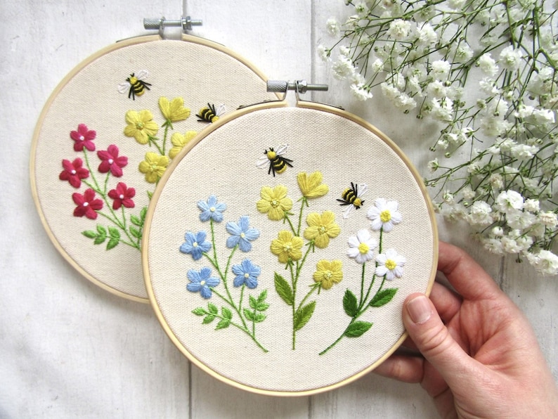 May include: Two round embroidery hoops with floral designs. One hoop features red flowers, the other blue and yellow flowers. Both have embroidered bees. The hoops are set against a white background with white flowers.