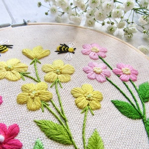 May include: Embroidered floral hoop art featuring yellow and pink flowers with green stems and leaves. Two embroidered bees are also included in the design. The artwork is set against a neutral fabric background, and framed in a wooden hoop.