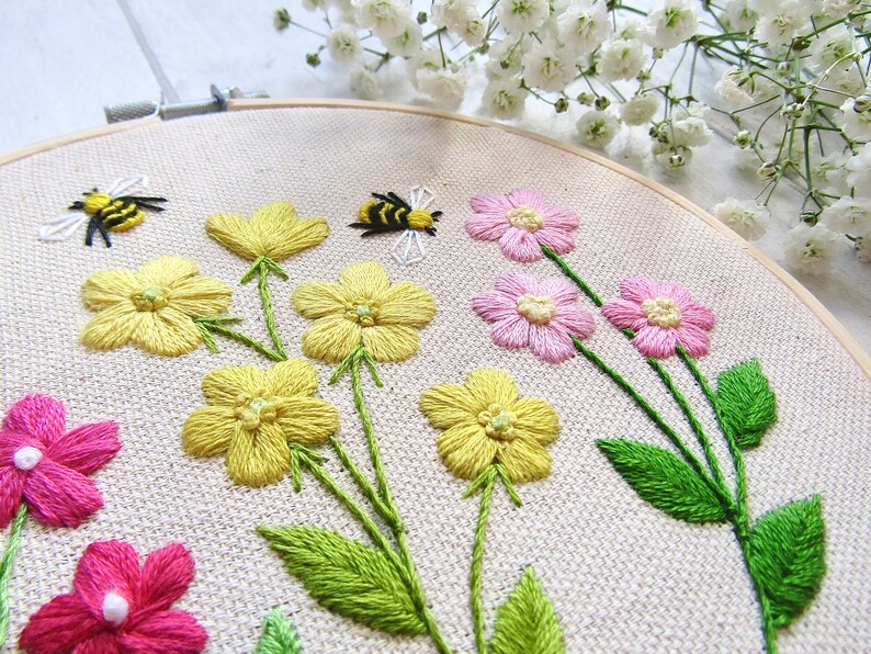 May include: Embroidered floral hoop art featuring yellow and pink flowers with green stems and leaves. Two embroidered bees are also included in the design. The artwork is set against a neutral fabric background, and framed in a wooden hoop.