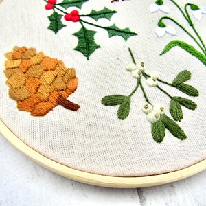 May include: Embroidered hoop art featuring botanical elements. A detailed pinecone in shades of brown and tan is displayed alongside sprigs of holly with red berries, mistletoe with white berries, and snowdrop flowers. The design is set against a neutral fabric.