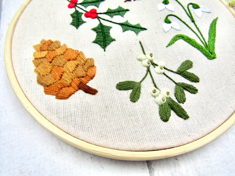May include: Embroidered hoop art featuring botanical elements. A detailed pinecone in shades of brown and tan is displayed alongside sprigs of holly with red berries, mistletoe with white berries, and snowdrop flowers. The design is set against a neutral fabric.