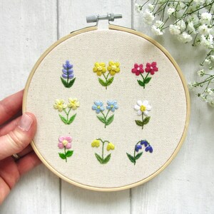 Op de afbeelding: Geborduurde bloemen op een borduurring. Het ontwerp toont verschillende bloemen in blauwe, gele, roze en witte tinten. De borduursels zijn geplaatst in een houten ring.