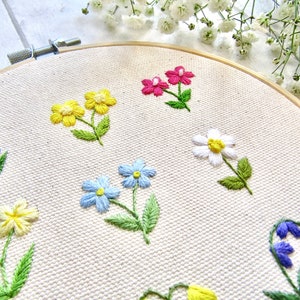 Op de afbeelding: Geborduurde bloemen op een cr&egrave;mekleurige stof, gespannen in een houten borduurring. De bloemen zijn in verschillende kleuren, waaronder geel, roze, blauw en wit, met groene bladeren. De borduursels zijn gedetailleerd en delicaat.