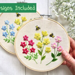 May include: Two embroidery hoops with floral designs. One hoop features blue flowers, the other pink and yellow flowers. Both have green leaves and a bee. The text "2 Designs Included" is at the top.