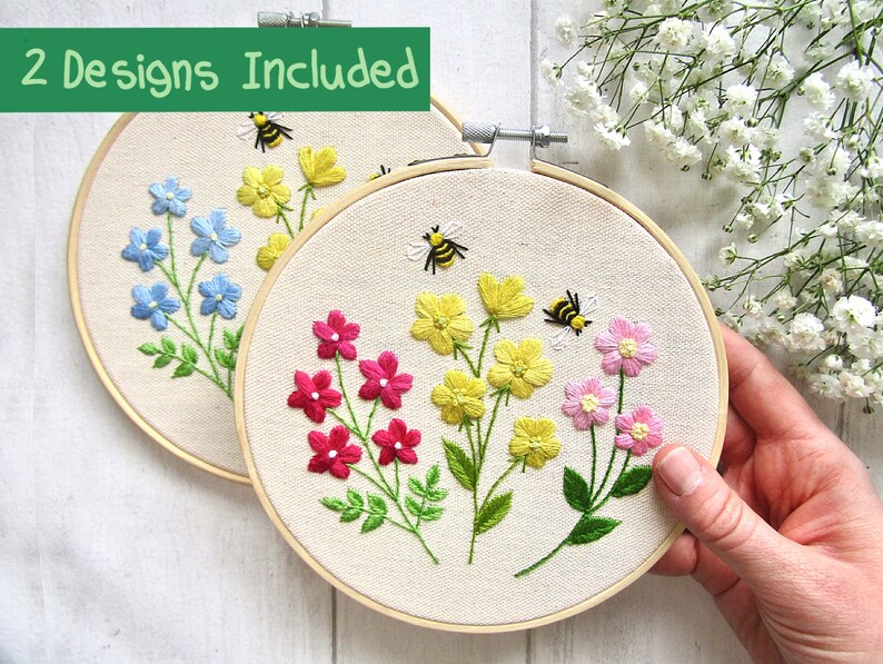 May include: Two embroidery hoops with floral designs. One hoop features blue flowers, the other pink and yellow flowers. Both have green leaves and a bee. The text "2 Designs Included" is at the top.