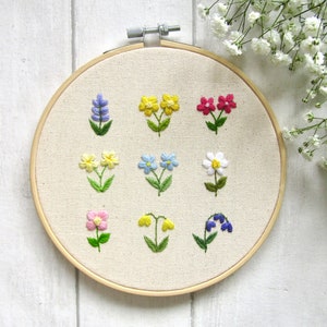 Op de afbeelding: Een handgeborduurde borduurring met negen verschillende kleurrijke bloemmotieven. De bloemen omvatten lavendel, geel, roze, blauw en wit met groene stelen en bladeren, alles tegen een neutrale stoffen achtergrond.