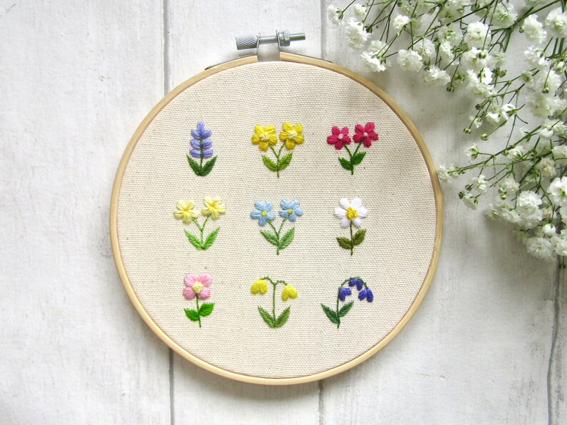 Op de afbeelding: Een handgeborduurde borduurring met negen verschillende kleurrijke bloemmotieven. De bloemen omvatten lavendel, geel, roze, blauw en wit met groene stelen en bladeren, alles tegen een neutrale stoffen achtergrond.