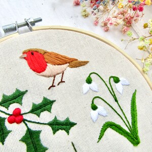 May include: Embroidered artwork in a wooden hoop featuring a robin with a red breast, holly with red berries, and snowdrop flowers. The embroidery is on a cream-colored fabric, with a wooden hoop and a white background.