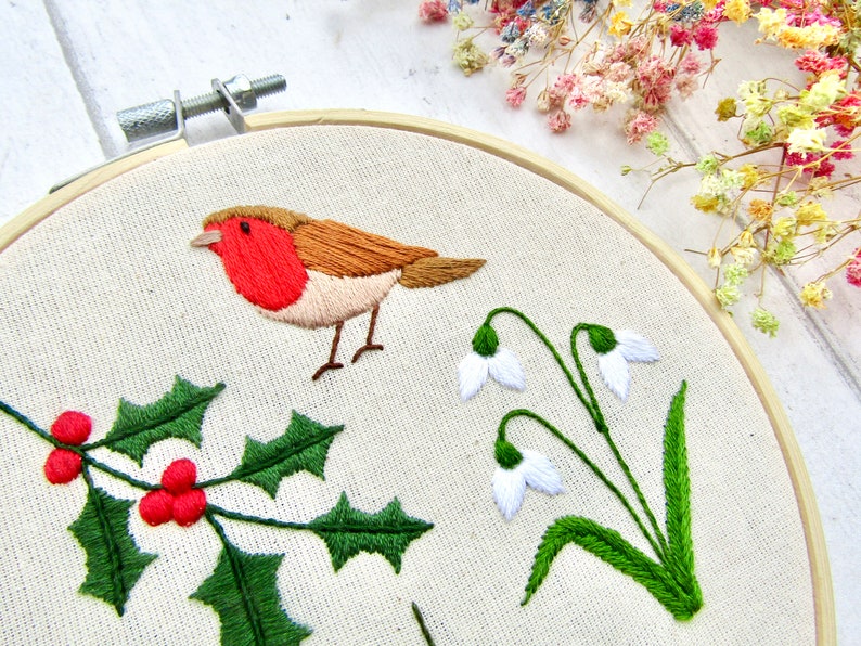 May include: Embroidered artwork in a wooden hoop featuring a robin with a red breast, holly with red berries, and snowdrop flowers. The embroidery is on a cream-colored fabric, with a wooden hoop and a white background.