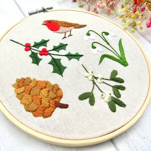 May include: Embroidered hoop art featuring a robin, holly with red berries, snowdrops, a pine cone, and mistletoe. The embroidery is done on a cream-colored fabric, set in a wooden hoop. The design evokes a winter or holiday theme.
