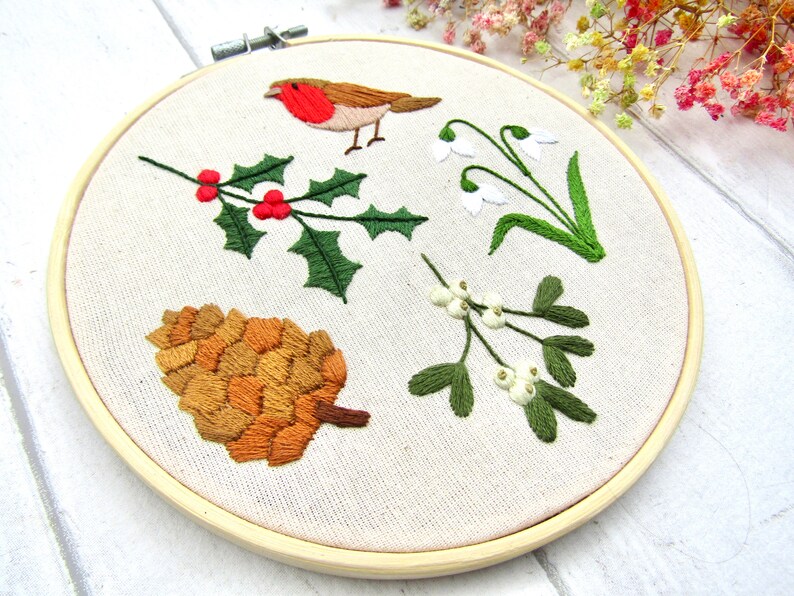 May include: Embroidered hoop art featuring a robin, holly with red berries, snowdrops, a pine cone, and mistletoe. The embroidery is done on a cream-colored fabric, set in a wooden hoop. The design evokes a winter or holiday theme.