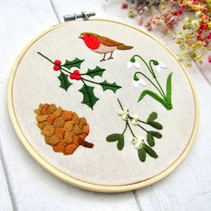May include: Embroidered hoop art featuring a robin, holly with red berries, snowdrops, a pine cone, and mistletoe. The embroidery is done on a beige fabric, set in a wooden hoop. The design has a seasonal, nature-inspired theme.