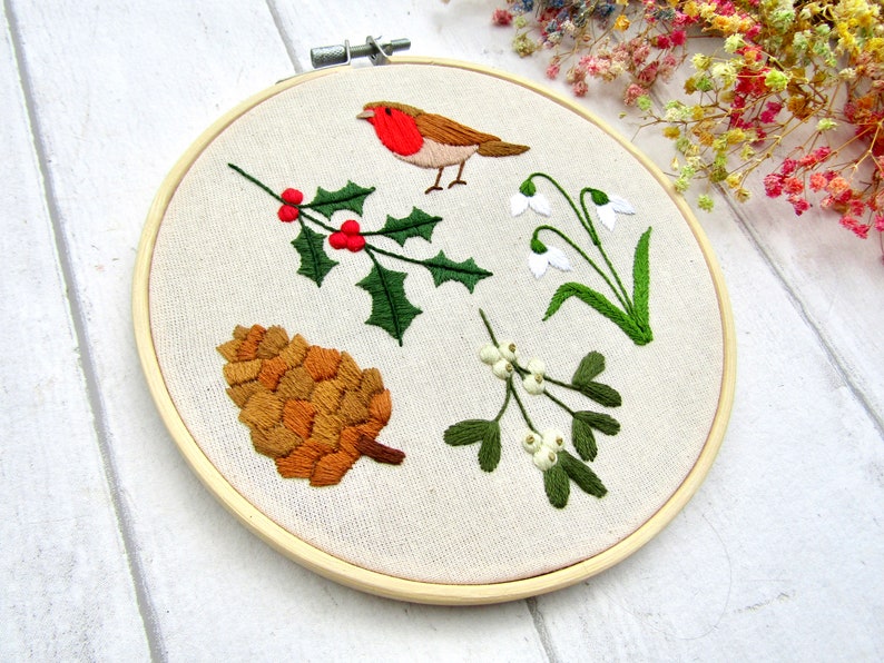 May include: Embroidered hoop art featuring a robin, holly with red berries, snowdrops, a pine cone, and mistletoe. The embroidery is done on a beige fabric, set in a wooden hoop. The design has a seasonal, nature-inspired theme.