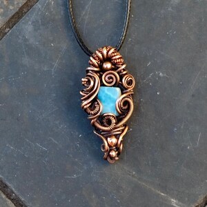 May include: Copper wire-wrapped pendant with a light blue gemstone, suspended from a black cord. The pendant features intricate wirework in a swirling pattern, showcasing the gemstone's color and shape. A unique handcrafted necklace.