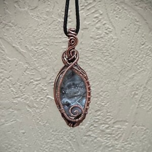 May include: A handcrafted pendant featuring a gray stone encased in copper wire. The wire is intricately woven around the stone, forming a decorative frame and bail. The pendant hangs from a black cord, showcasing a unique, artistic design.