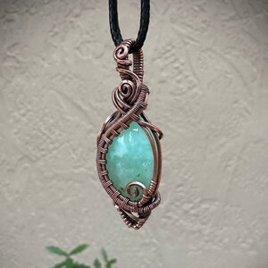 May include: A handcrafted pendant featuring a light green gemstone wrapped in copper wire. The wire is intricately woven around the stone, creating a decorative cage. The pendant hangs from a black cord, showcasing the artisan's skill.