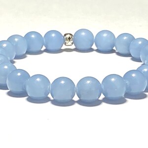 May include: A light blue beaded bracelet. The bracelet features round, smooth beads in a soft, pastel blue color. A small, silver-colored spacer bead with a decorative pattern is centered on the bracelet. The beads are strung together to form a complete circle.