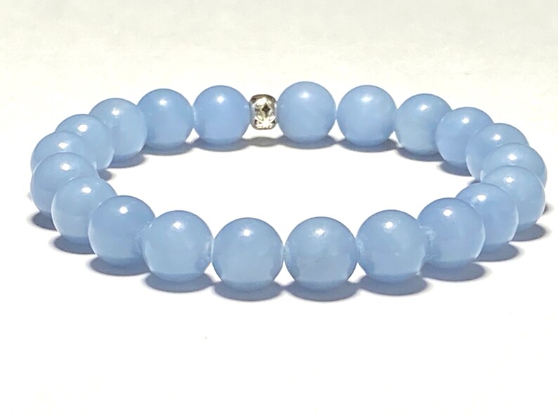 May include: A light blue beaded bracelet. The bracelet features round, smooth beads in a soft, pastel blue color. A small, silver-colored spacer bead with a decorative pattern is centered on the bracelet. The beads are strung together to form a complete circle.