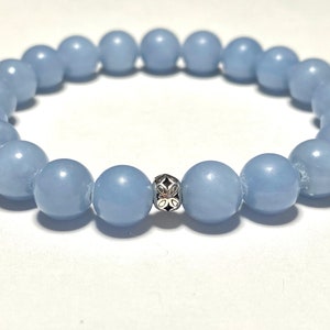 May include: A light blue beaded bracelet with a silver accent bead. The bracelet is made of round, smooth beads. The silver bead has a decorative floral design. The bracelet is on a white background.