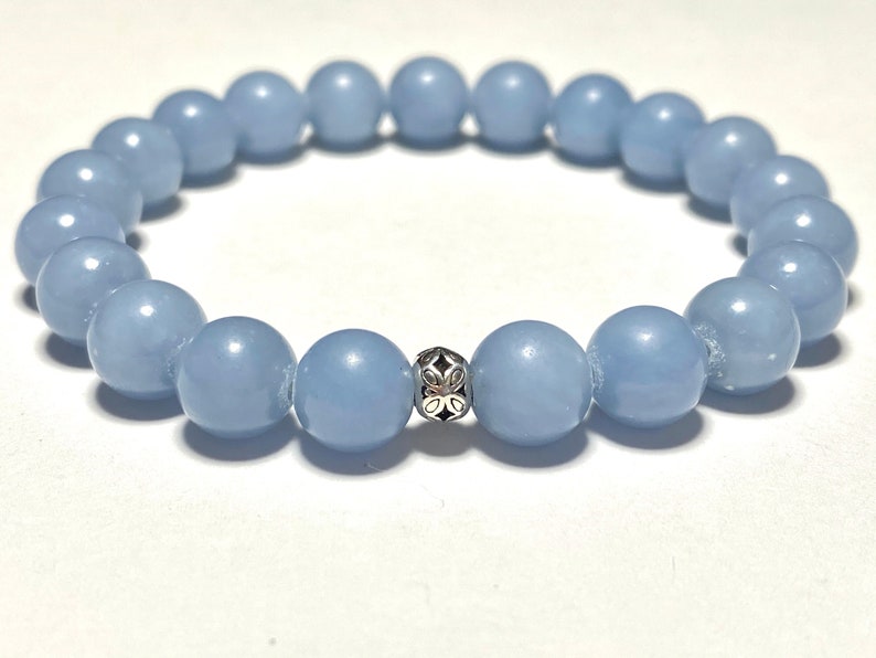 May include: A light blue beaded bracelet with a silver accent bead. The bracelet is made of round, smooth beads. The silver bead has a decorative floral design. The bracelet is on a white background.