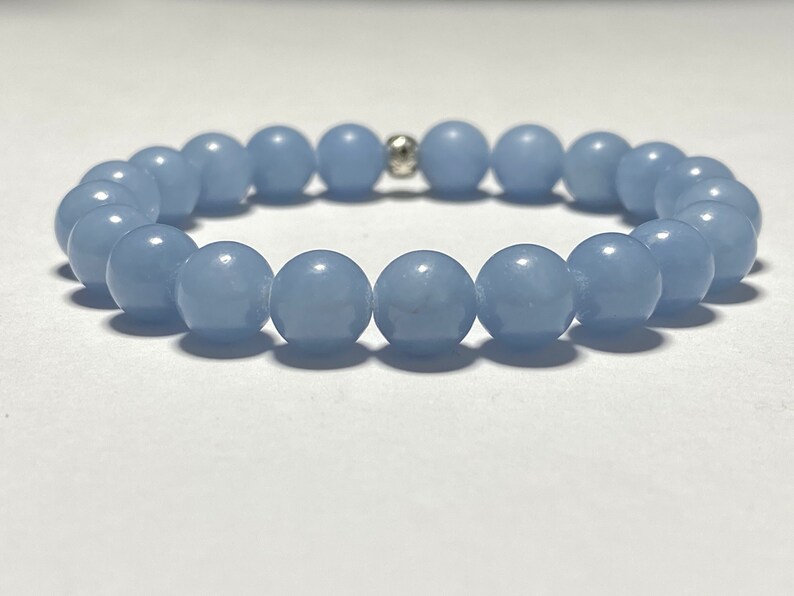 May include: A light blue beaded bracelet. The bracelet features round, smooth beads strung together with a silver-colored accent bead. The beads are a soft, pastel blue color, and the bracelet is displayed on a white surface.