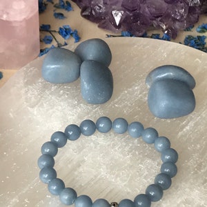 May include: A light blue beaded bracelet with a silver accent bead is displayed on a white surface, alongside three blue stones. A large amethyst cluster and a pink crystal are in the background.