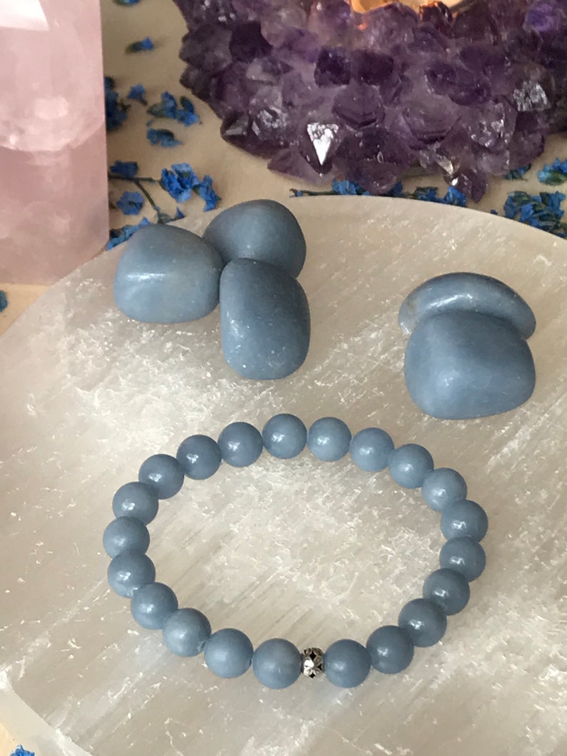 May include: A light blue beaded bracelet with a silver accent bead is displayed on a white surface, alongside three blue stones. A large amethyst cluster and a pink crystal are in the background.