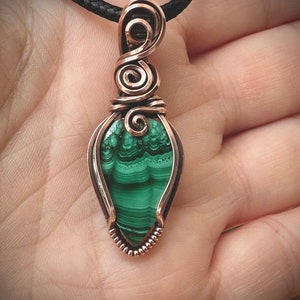 May include: A handcrafted copper wire pendant with a teardrop-shaped malachite stone. The green stone has dark green and black patterns. The pendant is suspended from a black cord necklace. The copper wire is intricately wrapped around the stone.